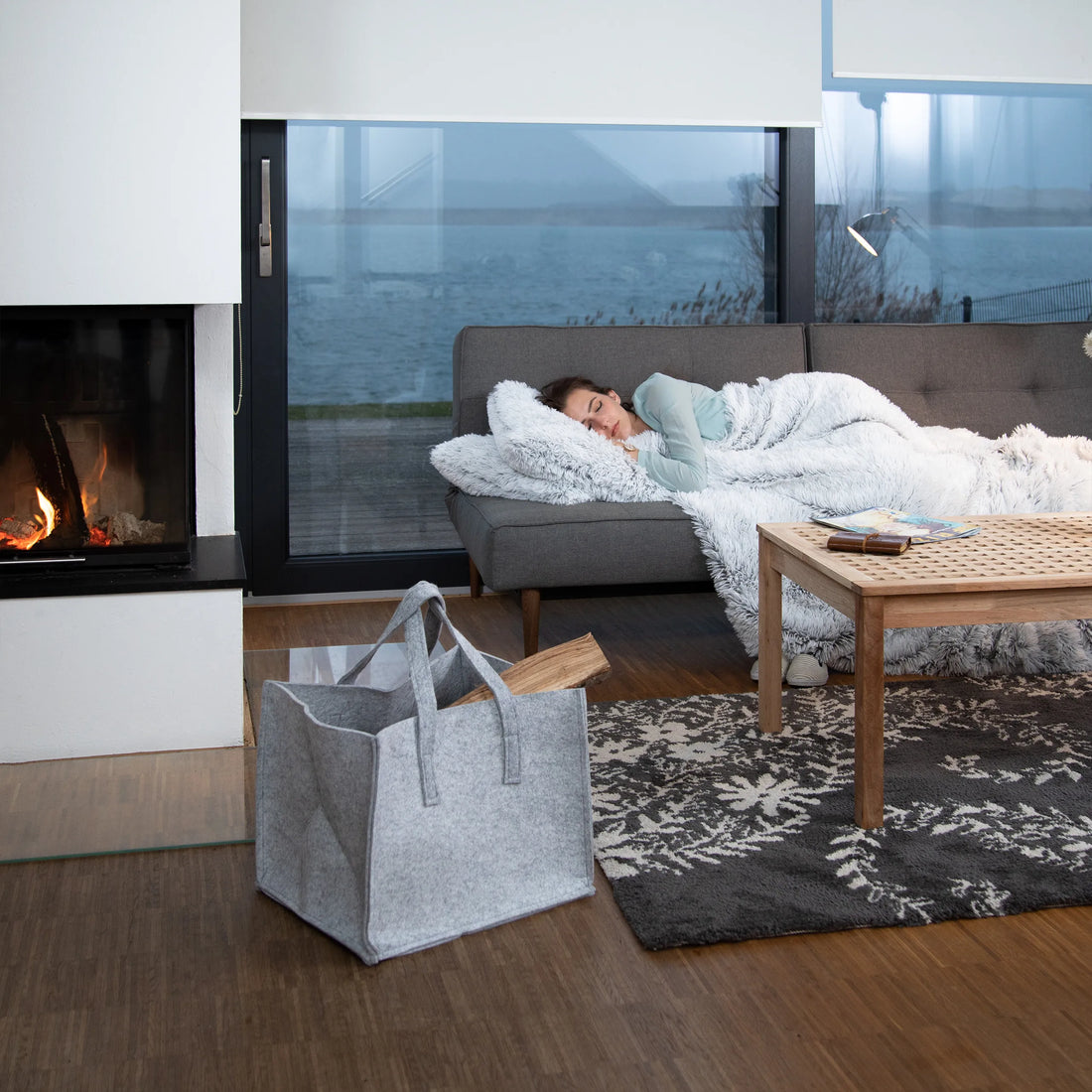 Filz-Holzkorb mit Brennholz auf dem Teppich im Wohnzimmer, im Hintergrund Sofa und Kamin