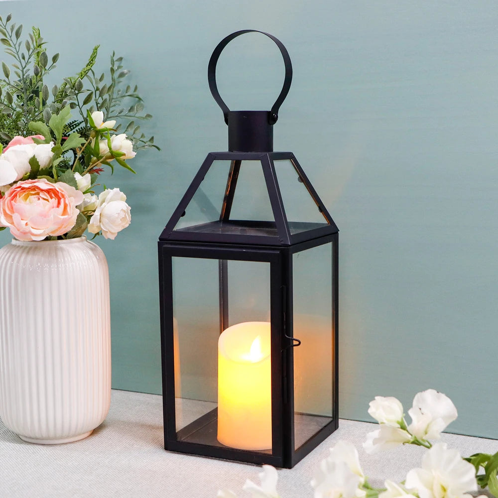 Schwarzes Windlicht mit flackernder Kerze neben weißer Vase mit Rosen