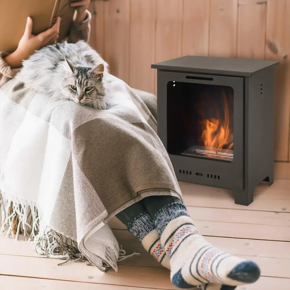 Schwarzer Ethanol-Standkamin auf Holzboden, davor Beine in Wollsocken unter Decke mit Katze